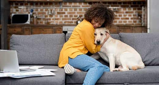 Mujer con un jersey amarillo abrazando a su labrador amarillo sentada en un sofá gris.